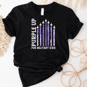 May include: Black t-shirt with a purple up for military kids design. The design features a purple and white American flag with airplanes flying over it.