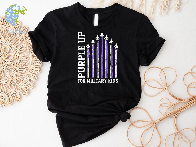 May include: Black t-shirt with a purple up for military kids design. The design features a purple and white American flag with airplanes flying over it.