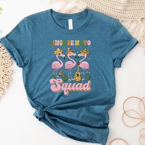 以下が含まれることがあります： ターコイズブルーのTシャツに、ソンブレロをかぶってマラカスとギターを持ったピンクのフラミンゴ3羽のグラフィックがプリントされています。フラミンゴの下には「Cinco de Mayo Squad」の文字がプリントされています。