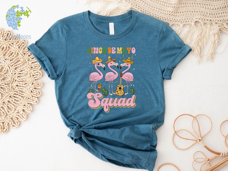以下が含まれることがあります： ターコイズブルーのTシャツに、ソンブレロをかぶってマラカスとギターを持ったピンクのフラミンゴ3羽のグラフィックがプリントされています。フラミンゴの下には「Cinco de Mayo Squad」の文字がプリントされています。