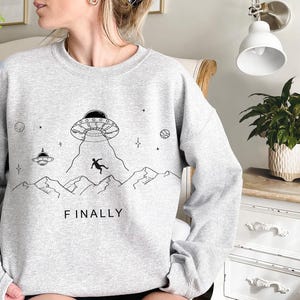 K&ouml;nnte beinhalten: Ein graues Sweatshirt mit einem Aufdruck eines UFOs und einer Person, die daraus f&auml;llt. Der Aufdruck ist von Sternen umgeben und der Text "FINALLY" ist darunter gedruckt.