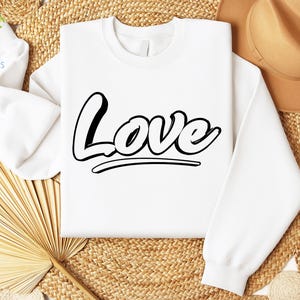 May include: White sweatshirt with the word "Love" printed in black, stylized lettering. The sweatshirt is laying on a woven straw surface.