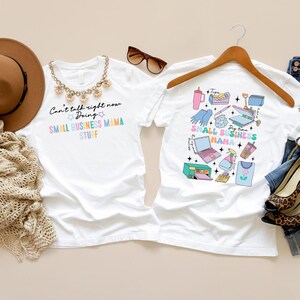 K&ouml;nnte beinhalten: Zwei wei&szlig;e T-Shirts mit einem kleinen Gesch&auml;ftsmotiv. Ein Shirt tr&auml;gt den Text "Can't talk right now Doing SMALL BUSINESS MAMA STUFF". Das andere Shirt zeigt eine bunte Grafik mit kleinen Gesch&auml;ftsutensilien, darunter ein Laptop, ein Drucker, eine Hei&szlig;presse und eine Tasse Kaffee.
