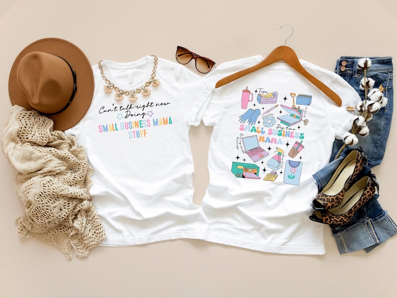K&ouml;nnte beinhalten: Zwei wei&szlig;e T-Shirts mit einem kleinen Gesch&auml;ftsmotiv. Ein Shirt tr&auml;gt den Text "Can't talk right now Doing SMALL BUSINESS MAMA STUFF". Das andere Shirt zeigt eine bunte Grafik mit kleinen Gesch&auml;ftsutensilien, darunter ein Laptop, ein Drucker, eine Hei&szlig;presse und eine Tasse Kaffee.
