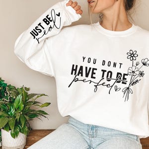 K&ouml;nnte beinhalten: Wei&szlig;er Langarm-Sweatshirt mit dem Text "Just Be Real" auf dem &Auml;rmel und "You Don't Have To Be Perfect" auf der Vorderseite mit einem kleinen Herzen und einem Blumenstrau&szlig;.