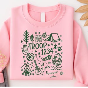 May include: A light pink sweatshirt with a green camping-themed graphic. The design includes trees, a tent, a map, a compass, a campfire, a guitar, and the text "TROOP 1234" and "Harper".