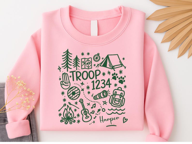 May include: A light pink sweatshirt with a green camping-themed graphic. The design includes trees, a tent, a map, a compass, a campfire, a guitar, and the text "TROOP 1234" and "Harper".