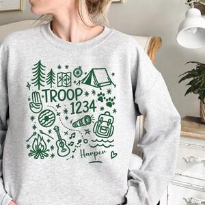May include: A light gray sweatshirt with a green camping-themed graphic. The design includes trees, a tent, a map, a compass, a guitar, a campfire, and the text "TROOP 1234". The name "Harper" is also included.