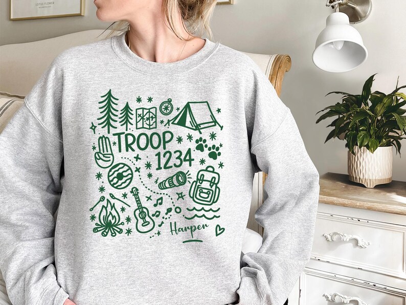 May include: A light gray sweatshirt with a green camping-themed graphic. The design includes trees, a tent, a map, a compass, a guitar, a campfire, and the text "TROOP 1234". The name "Harper" is also included.