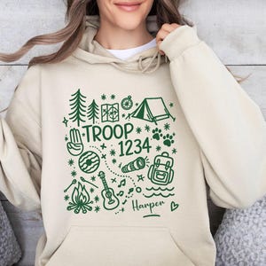 May include: Beige hoodie with a green graphic design featuring camping-themed illustrations, including trees, a tent, and a guitar. The text "TROOP 1234" is prominently displayed, with the name "Harper" below. A cozy and casual garment.