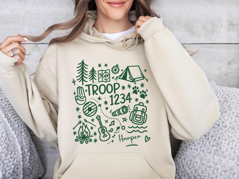 May include: Beige hoodie with a green graphic design featuring camping-themed illustrations, including trees, a tent, and a guitar. The text "TROOP 1234" is prominently displayed, with the name "Harper" below. A cozy and casual garment.
