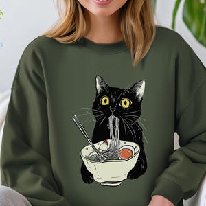 Peut inclure: Un chat noir aux yeux jaunes mange des nouilles ramen dans un bol blanc. Le chat porte un sweat-shirt vert avec un motif de chat mangeant des nouilles ramen.