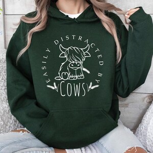 May include: A dark green hoodie with a white graphic that says "Easily Distracted By Cows" and features a cartoon cow with a heart on its chest.