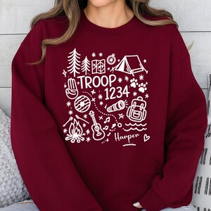 May include: Burgundy sweatshirt with a white camping-themed graphic. The design includes a tent, trees, map, compass, paw prints, and the text "TROOP 1234" and "Harper". A cozy, casual top for outdoor enthusiasts.