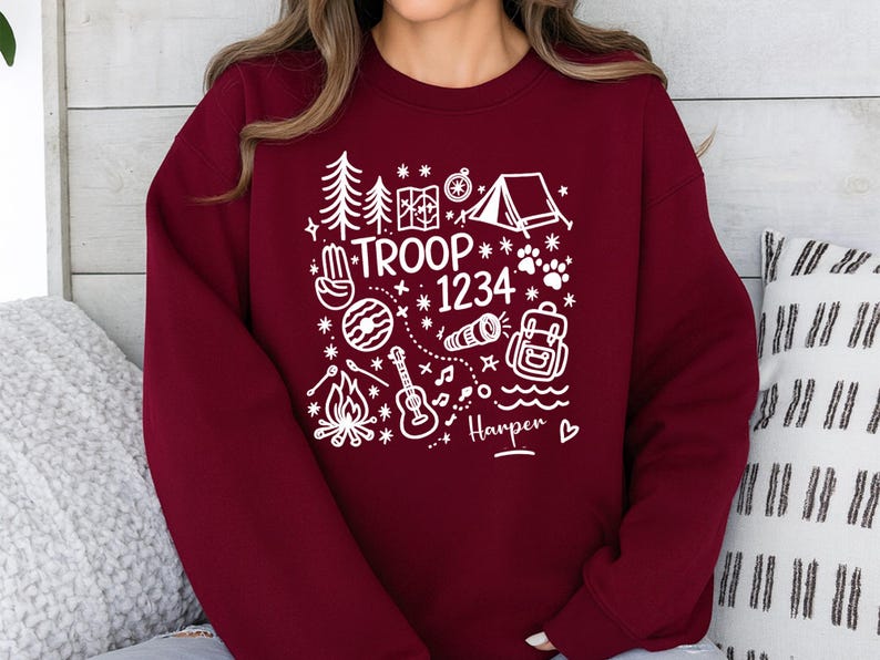 May include: Burgundy sweatshirt with a white camping-themed graphic. The design includes a tent, trees, map, compass, paw prints, and the text "TROOP 1234" and "Harper". A cozy, casual top for outdoor enthusiasts.