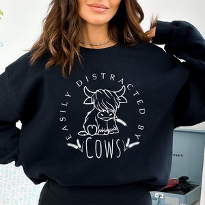 May include: Black sweatshirt with a white graphic of a Highland cow and the text "Easily Distracted By Cows".