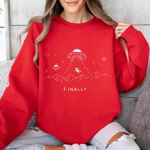 K&ouml;nnte beinhalten: Roter Pullover mit einem wei&szlig;en Aufdruck eines UFOs, einer Person, die aus dem UFO f&auml;llt, und dem Text "FINALLY".