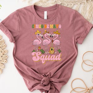以下が含まれることがあります： 「Cinco de Mayo Squad」という文字と、ソンブレロをかぶってギターを弾いているピンクのフラミンゴ3羽のグラフィックが描かれたピンクのTシャツです。