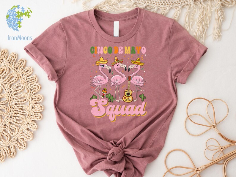 以下が含まれることがあります： 「Cinco de Mayo Squad」という文字と、ソンブレロをかぶってギターを弾いているピンクのフラミンゴ3羽のグラフィックが描かれたピンクのTシャツです。