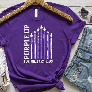 May include: Purple t-shirt with a white graphic of an American flag with airplanes and the text "Purple Up for Military Kids".