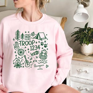 May include: A light pink sweatshirt with a green camping-themed graphic. The design includes trees, a tent, a compass, paw prints, and the words "TROOP 1234" with the name "Harper" below. The sweatshirt is a crew neck style.
