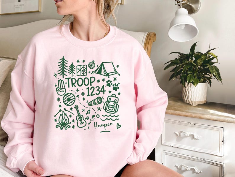 May include: A light pink sweatshirt with a green camping-themed graphic. The design includes trees, a tent, a compass, paw prints, and the words "TROOP 1234" with the name "Harper" below. The sweatshirt is a crew neck style.