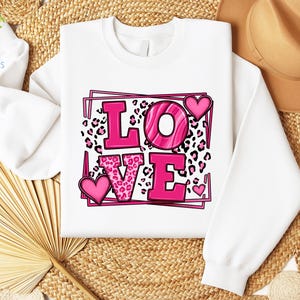 May include: White sweatshirt with a pink "LOVE" graphic. The letters are outlined in black and have a pink leopard print pattern. There are pink hearts around the letters.