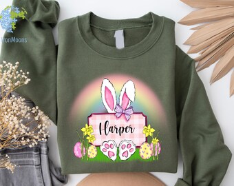 May include: A green sweatshirt with a pink and white bunny rabbit holding a sign that says "Harper". The bunny is in front of a rainbow and surrounded by Easter eggs and flowers.