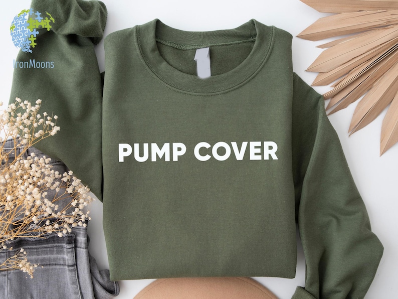 Pode incluir: Um moletom verde-oliva com as palavras "PUMP COVER" impressas em branco. O moletom est&aacute; dobrado, com as mangas vis&iacute;veis. O fundo inclui plantas secas e um par de jeans.