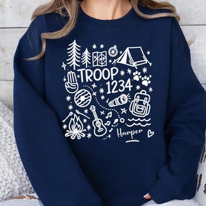 May include: Navy blue sweatshirt with a white graphic design featuring camping-themed illustrations and the text "TROOP 1234" along with a name. The design includes a tent, trees, a compass, and a guitar.