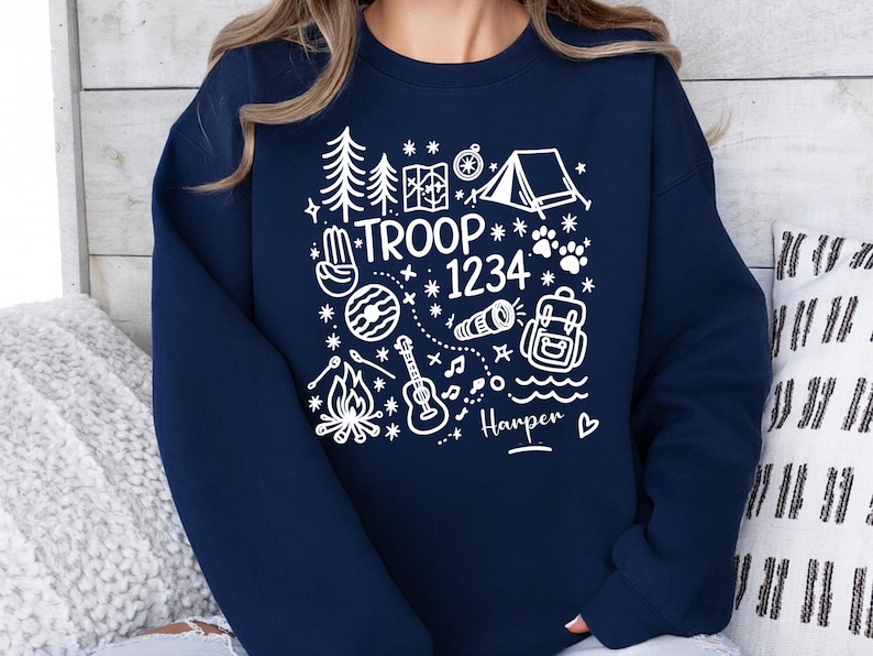 May include: Navy blue sweatshirt with a white graphic design featuring camping-themed illustrations and the text "TROOP 1234" along with a name. The design includes a tent, trees, a compass, and a guitar.