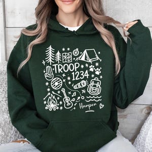 May include: Dark green hoodie with white graphic illustrations of camping elements, including trees, a tent, and a campfire. The text "TROOP 1234" is displayed, along with a name and a heart symbol. The hoodie has a hood and a front pocket.