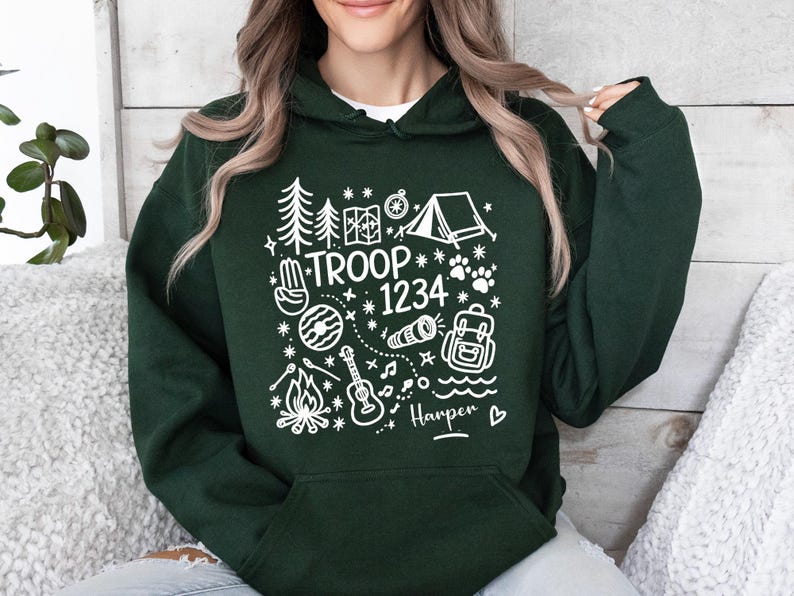 May include: Dark green hoodie with white graphic illustrations of camping elements, including trees, a tent, and a campfire. The text "TROOP 1234" is displayed, along with a name and a heart symbol. The hoodie has a hood and a front pocket.