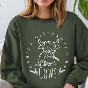 May include: A dark green sweatshirt with a white graphic design. The graphic features a cartoon highland cow with the text "Easily Distracted By Cows".