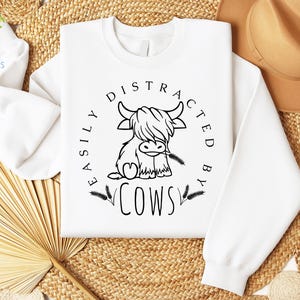 May include: White sweatshirt with a black graphic of a Highland cow and the text "Easily Distracted By Cows".
