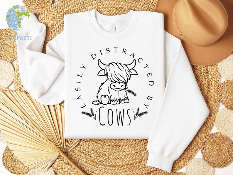 May include: White sweatshirt with a black graphic of a Highland cow and the text "Easily Distracted By Cows".