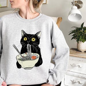 Peut inclure: Un sweat-shirt gris avec un chat noir mangeant des nouilles ramen dans un bol. Le chat a les yeux jaunes et un bavoir blanc.