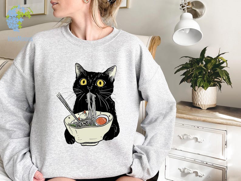 Peut inclure: Un sweat-shirt gris avec un chat noir mangeant des nouilles ramen dans un bol. Le chat a les yeux jaunes et un bavoir blanc.