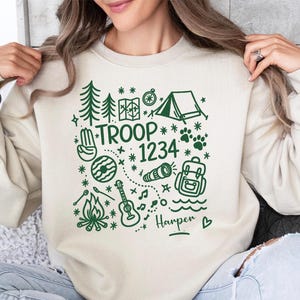 May include: Cream-colored sweatshirt with a green graphic design featuring camping and outdoor adventure elements. The design includes trees, a tent, a compass, and the text "TROOP 1234" with a name below.
