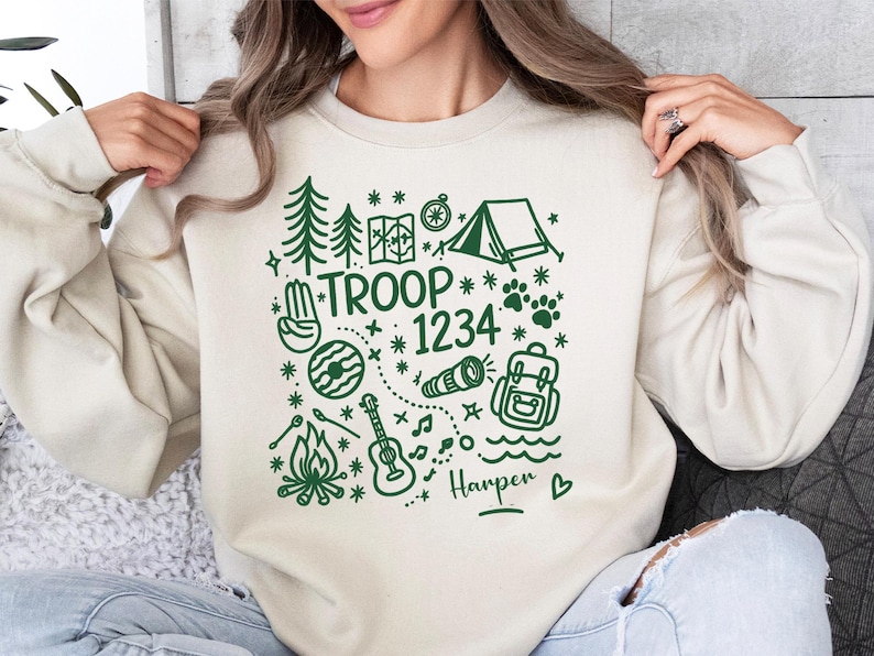 May include: Cream-colored sweatshirt with a green graphic design featuring camping and outdoor adventure elements. The design includes trees, a tent, a compass, and the text "TROOP 1234" with a name below.
