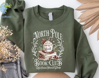May include: Olive green sweatshirt with a graphic that reads "North Pole Book Club." The design features a mug of hot cocoa, gingerbread man, candy cane, and books. The words "Hot Cocoa Served Daily" are also included.