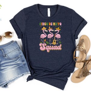 以下が含まれることがあります： 紺色のTシャツに、ソンブレロをかぶってギターを持ったピンクのフラミンゴ3羽のグラフィックがプリントされています。フラミンゴの下には「Cinco de Mayo Squad」という文字がプリントされています。