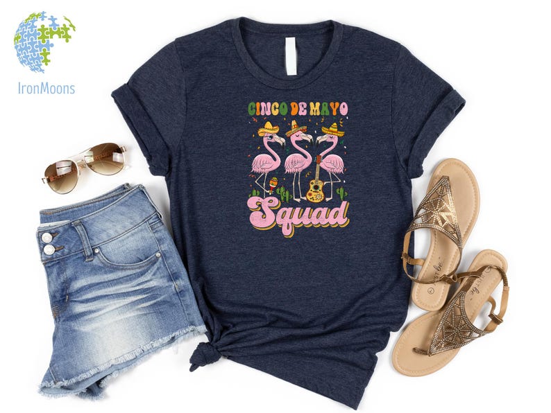 以下が含まれることがあります： 紺色のTシャツに、ソンブレロをかぶってギターを持ったピンクのフラミンゴ3羽のグラフィックがプリントされています。フラミンゴの下には「Cinco de Mayo Squad」という文字がプリントされています。
