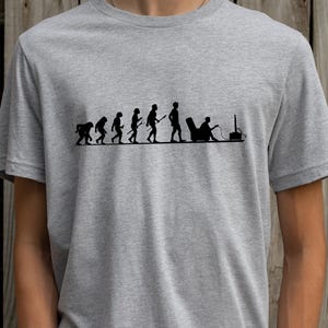 May include: A light gray t-shirt featuring a black graphic depicting the evolution of man, culminating in a person seated in a chair playing a video game. The design shows a progression from ape to gamer.