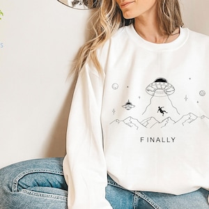K&ouml;nnte beinhalten: Wei&szlig;er Pullover mit einem schwarz-wei&szlig;en Grafikdruck eines UFOs, Bergen und einer Person, die vom Himmel f&auml;llt. Der Text "FINALLY" ist unter dem Grafikdruck aufgedruckt.