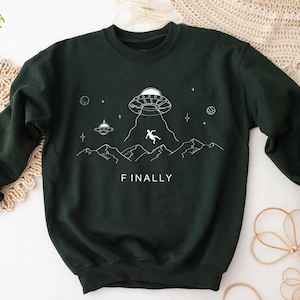 K&ouml;nnte beinhalten: Ein dunkelgr&uuml;ner Pullover mit einem wei&szlig;en Aufdruck eines UFOs, einer Person, die vom Himmel f&auml;llt, und Bergen. Der Text "FINALLY" ist unter dem Aufdruck gedruckt.