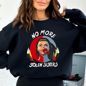 May include: Black sweatshirt featuring a graphic with the text "NO MORE STOLEN SISTERS." The design includes a woman's face, split between red and blue, with a handprint over her mouth. Feathers frame the image.