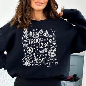 May include: Black sweatshirt with a white graphic design featuring camping-themed illustrations, the text "TROOP 1234", and the name "Harper". The design includes trees, a tent, a compass, a campfire, and a guitar.