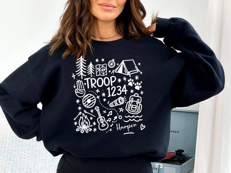 May include: Black sweatshirt with a white graphic design featuring camping-themed illustrations, the text "TROOP 1234", and the name "Harper". The design includes trees, a tent, a compass, a campfire, and a guitar.
