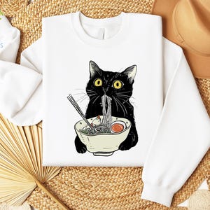 Peut inclure: Sweat-shirt blanc avec un chat noir mangeant des nouilles ramen dans un bol. Le chat a les yeux jaunes et un nez noir.